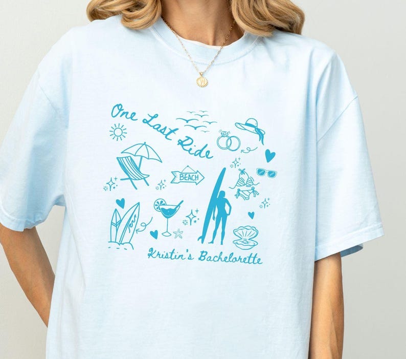 May include: Light blue t-shirt with a blue graphic design of a beach scene with the text "One Last Ride" and "Kristin's Bachelorette".