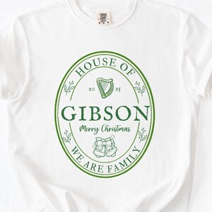 May include: White t-shirt with a green oval design. The design includes the words "HOUSE OF GIBSON" and "WE ARE FAMILY" in a circular pattern. A harp and bells are also featured, with "Merry Christmas" in the center. A festive holiday tee.