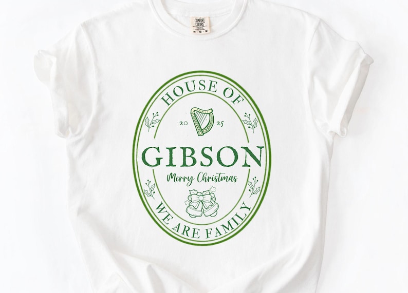 May include: White t-shirt with a green oval design. The design includes the words "HOUSE OF GIBSON" and "WE ARE FAMILY" in a circular pattern. A harp and bells are also featured, with "Merry Christmas" in the center. A festive holiday tee.