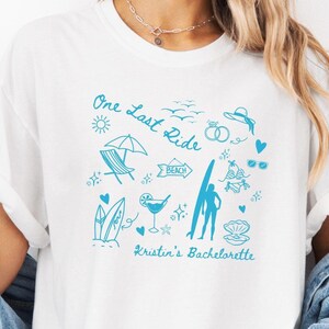 May include: White t-shirt with a blue graphic design that says "One Last Ride" and includes beach-themed illustrations like a surfboard, palm trees, sunglasses, and a cocktail. The text "Kristin's Bachelorette" is also included in the design.