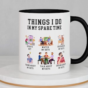 May include: White ceramic mug with a black handle and rim, featuring the text "THINGS I DO IN MY SPARE TIME." The mug has illustrations depicting activities like sports betting, watching games, and researching bets. The mug is on a white pedestal.
