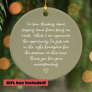 May include: A round, olive-green ceramic ornament with a gold string for hanging. The ornament has white text that reads, "I've been thinking about stepping down from being an adult..." A red box at the bottom says "Gift box included!"