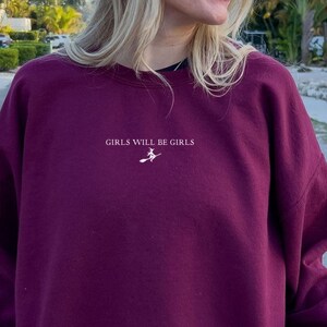 May include: A burgundy sweatshirt with the text "GIRLS WILL BE GIRLS" in white, along with a small graphic of a witch on a broom. The sweatshirt is a solid color and has a crew neck.