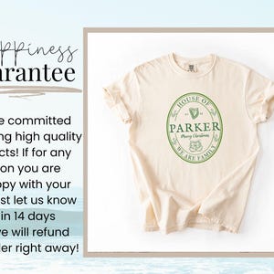 May include: Cream-colored t-shirt with a green oval design that reads "HOUSE OF PARKER Merry Christmas WE ARE FAMILY". The shirt has rolled-up sleeves and is displayed on a white background.