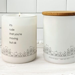 May include: Two white soy wax candles with a town silhouette design. One candle is lit, displaying the text "it's rude that you're moving but ok." The other candle has a wooden lid and the text "HAND POURED / 100% SOY WAX / 10 oz."