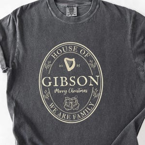 May include: Dark gray t-shirt with a cream-colored oval design. The design includes the text "HOUSE OF GIBSON" above "Merry Christmas" and "WE ARE FAMILY". A harp and bells are also featured.