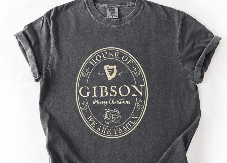 May include: Dark gray t-shirt with a cream-colored oval design. The design includes the text "HOUSE OF GIBSON" above "Merry Christmas" and "WE ARE FAMILY". A harp and bells are also featured.