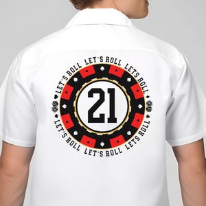 May include: White button-up shirt with a large graphic on the back. The graphic is a casino chip design with the number 21 in the center, surrounded by the words "LET'S ROLL" and card suit symbols. The shirt has short sleeves.