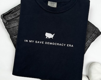 Puede incluir: Camiseta negra con un contorno blanco de los Estados Unidos y el texto "In My Save Democracy Era".