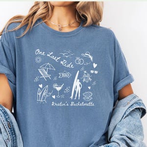 May include: A light blue t-shirt with a white graphic design featuring a surfer, beach umbrella, and the text "One Last Ride Kristin's Bachelorette".