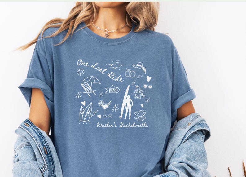 May include: A light blue t-shirt with a white graphic design featuring a surfer, beach umbrella, and the text "One Last Ride Kristin's Bachelorette".