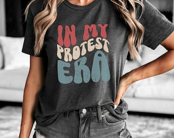 Op de afbeelding: Donkergrijs t-shirt met de tekst "IN MY PROTEST ERA" in een retro lettertype. De tekst is in rood, beige en blauw. Het shirt heeft een ronde hals en korte mouwen.