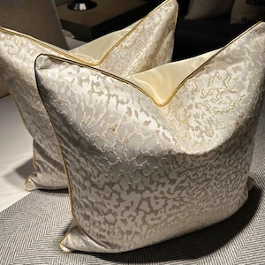 Op de afbeelding: Twee decoratieve kussens met een crèmekleurige achtergrond en een goudkleurig patroon. De kussens hebben een gouden rand en liggen op een grijs en wit visgraatpatroon dekbed. Deze kussens voegen luxe toe aan elke slaapkamer.