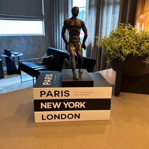 Può includere: Una scultura color bronzo di un uomo in posizione di camminata, sopra una pila di tre libri decorativi. I libri sono bianchi con testo nero che recita "PARIS", "NEW YORK" e "LONDON". Il libro in alto recita anche "CITY OF FASHION AND ROMANCE".