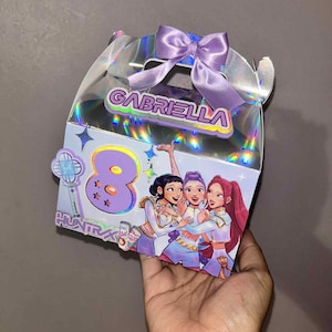 May include: A clear, iridescent gift box with a purple bow and the name "GABRIELLA" in a holographic font. The box features a cartoon image of three girls and the number "8".
