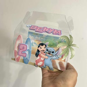 Puede incluir: Caja de plástico transparente con una escena tropical que presenta a los personajes Lilo y Stitch. La caja tiene un número 2 rosa y azul, tablas de surf y el nombre "DENYM" en rosa. También se ve una palmera.