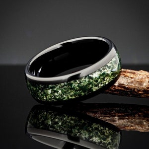 May include: A black ceramic ring with a green and white speckled inlay. The ring is sitting on a piece of wood.