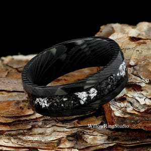 May include: Black carbon fiber ring with silver flecks embedded in the band. The ring is on a piece of tree bark.