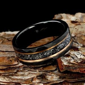 Peut inclure: Une bague noire présentant une surface extérieure à facettes avec une incrustation centrale de fragments métalliques sombres et texturés, bordée par deux fines bandes de couleur cuivre polies faisant le tour de l'anneau.