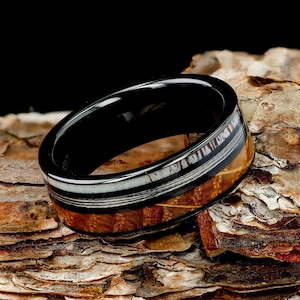 May include: A ring featuring a polished black band with a central inlay composed of a white textured material and a brown wood grain section. The ring has a smooth, reflective finish on the interior and exterior edges, showcasing a layered design.