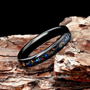 May include: A black ceramic ring with a thin band and a colorful inlay of blue, orange, and red flecks. The ring is sitting on a piece of wood.