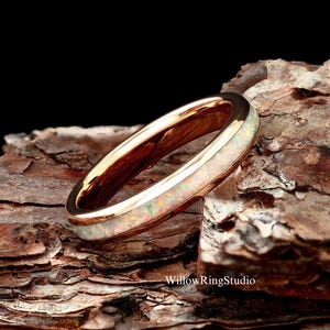 May include: A rose gold band ring featuring a central inlay of iridescent white opal with subtle flashes of green and pink. The ring has a polished, reflective metallic finish and is positioned at an angle. The text WillowRingStudio is visible.