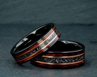 May include: Two rings featuring a black metal band with a central inlay of dark, textured material flanked by two thin copper-colored stripes. The rings have a polished finish and a flat profile, showcasing a contrast between the dark metal and the warm metallic accents.
