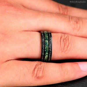Pu&ograve; includere: Anello nero con intarsio in pietra verde. L'anello ha una fascia nera con un design strutturato sui bordi. L'intarsio centrale &egrave; di un verde vibrante, creando un contrasto sorprendente. L'anello &egrave; mostrato su un dito.