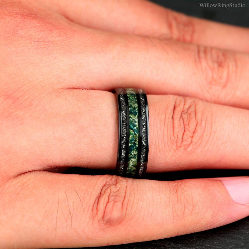 Pu&ograve; includere: Anello nero con intarsio in pietra verde. L'anello ha una fascia nera con un design strutturato sui bordi. L'intarsio centrale &egrave; di un verde vibrante, creando un contrasto sorprendente. L'anello &egrave; mostrato su un dito.