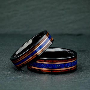 May include: Two rings with a black metal finish, featuring a central inlay of vibrant blue crushed stone flanked by two thin bands of reddish-brown wood. The rings have a smooth, polished surface and a flat profile, showcasing a layered design.