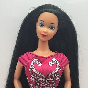 May include: A doll with long black hair and blue eyes. The doll is wearing a pink top with a white and floral design. The doll has red lipstick and small earrings. The doll's outfit includes a black and white patterned skirt.