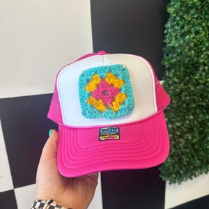 May include: A pink and white trucker hat with a crocheted square patch in turquoise, yellow, and pink. The patch is attached to the front of the hat. The hat has a mesh back and a label that says "OTTO WORLD FAMOUS TRUCKER".