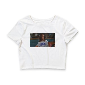 May include: A white short-sleeved crop top featuring a rectangular graphic print on the chest. The print displays a scene of a person with brown hair wearing a grey sweatshirt with the blue text KNICKS BASKETBALL printed across the front.