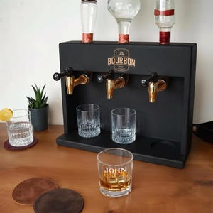 Könnte beinhalten: Ein mattschwarzer Spirituosenspender aus Holz mit drei goldfarbenen Metallhähnen und drei Flaschenhaltern auf der Oberseite. Die Vorderseite zeigt ein Logo mit dem Text BOURBON. Zwei klare Gläser stehen auf der Basis und ein Glas mit der Aufschrift JOHN davor.