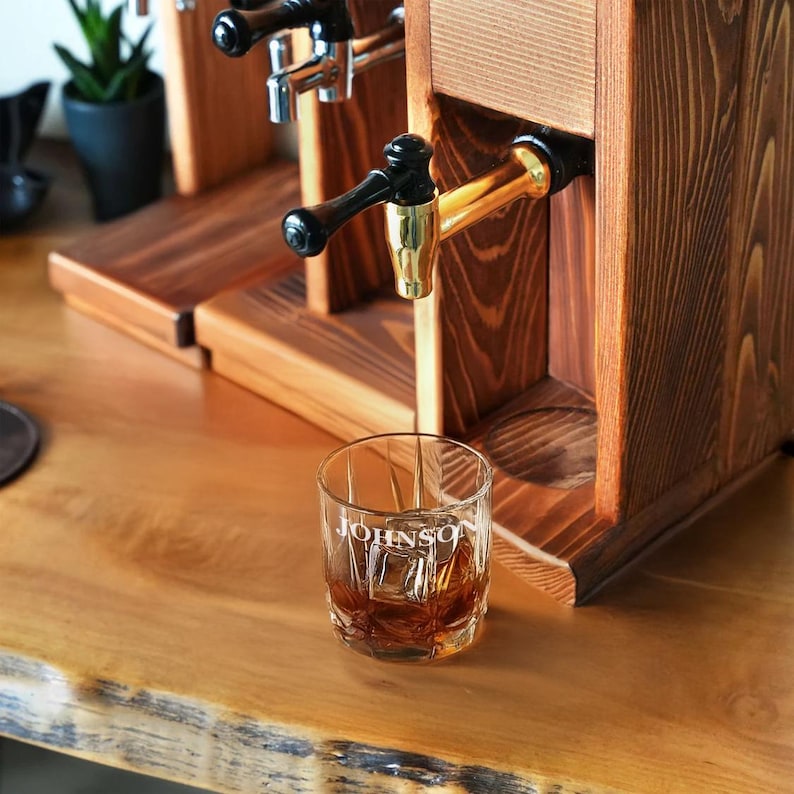 A wooden dispenser with a glass of whiskey in the foreground, and a potted plant in the background.