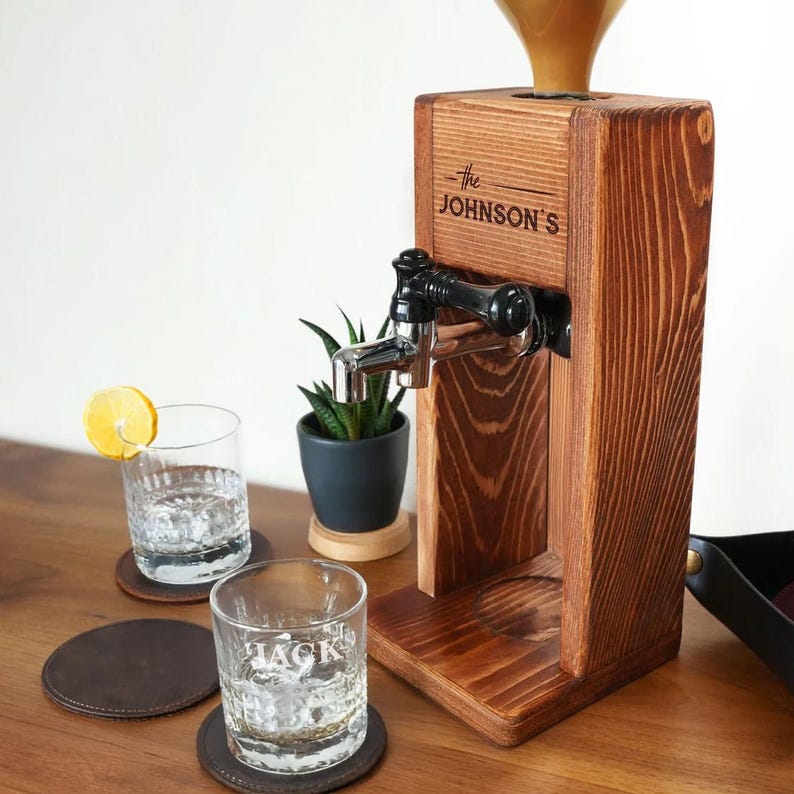 A wooden coffee dispenser with a black handle and spout, along with two glasses and a potted plant on a wooden table.
