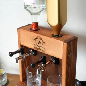A wooden tap dispenser with a gold-colored bottle and a clear glass bottle on top, along with a wooden shelf and a glass cup.