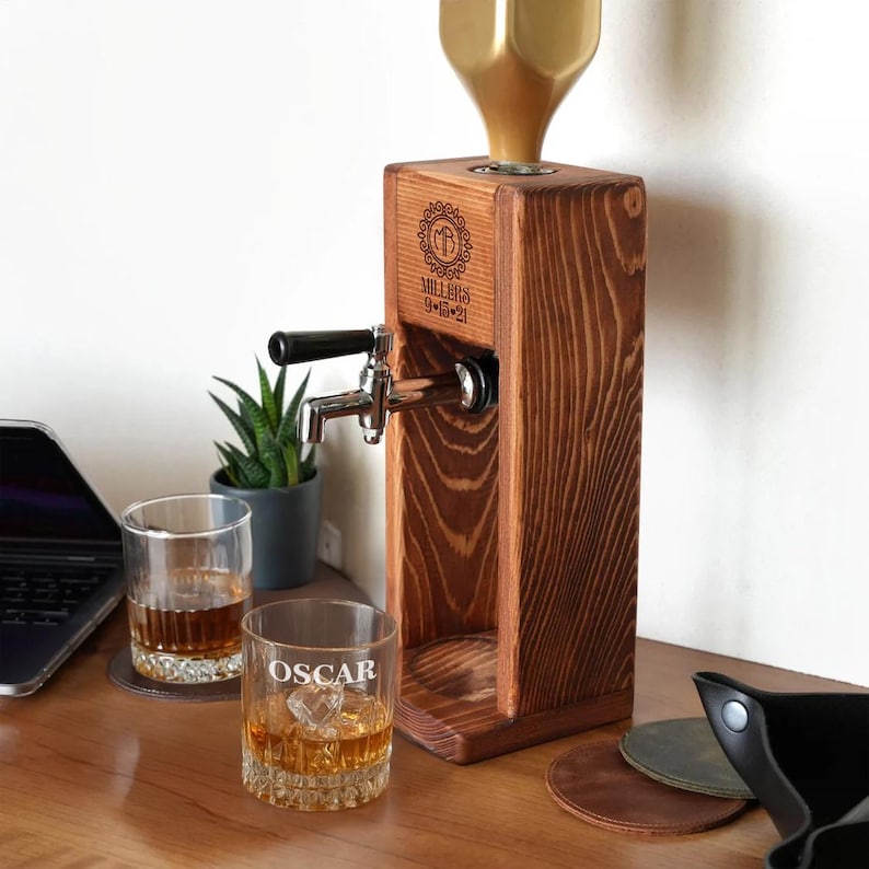 a wooden beer tap dispenser with a glass of amber-colored liquid, likely whiskey, placed on a wooden desk. There is also a laptop and a potted plant in the background.