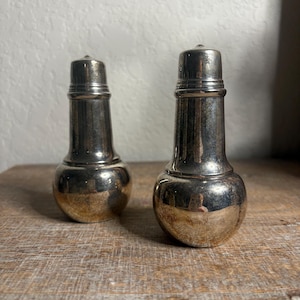 May include: Two antique silver salt and pepper shakers. The shakers have a bulbous base and a cylindrical top. The silver has a tarnished appearance, with a warm, golden hue at the base. They sit on a wooden surface.
