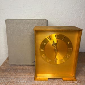 May include: A gold-coloured rectangular clock with Roman numerals on the face. The clock has a textured surface and is next to a grey box. The clock face is circular and the hands are black.