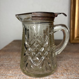 May include: A clear glass pitcher with a diamond pattern and a metal lid. The pitcher has a handle and a spout.