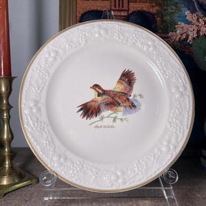 May include: White ceramic plate with a gold rim and a raised floral design. The center of the plate features a painted image of a bobwhite quail in flight with the text "Bob White" below.