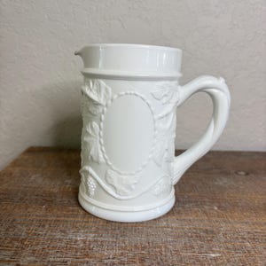Op de afbeelding: Een witte keramische kan met een handvat en een tuit. De kan is versierd met in reliëf aangebrachte wijnranken en bladeren rond een ovaal frame. De kan staat op een houten oppervlak.