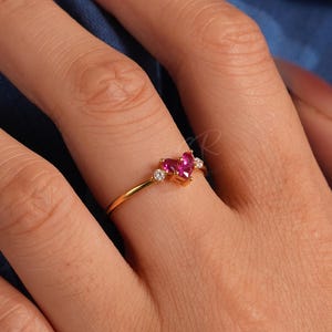 Puede incluir: Un anillo de oro con una piedra preciosa roja en forma de corazón, flanqueada por dos piedras claras más pequeñas. El anillo está engastado en una fina banda de oro. El anillo se muestra en una mano con uñas cuidadas, sobre un fondo azul.
