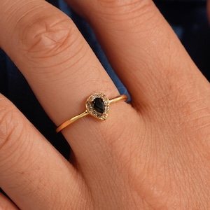 Puede incluir: Un anillo de oro con una piedra preciosa negra en forma de corazón. El anillo está engastado con pequeñas piedras transparentes alrededor del corazón. El anillo está en un dedo, sobre un fondo azul.