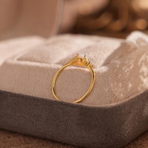 Puede incluir: Un anillo de oro con una gema rectangular blanquecina en el centro, flanqueada por piedras m&aacute;s peque&ntilde;as y transparentes. El anillo se presenta sobre una superficie gris y beige, mostrando su delicado dise&ntilde;o. La banda del anillo es fina y elegante.