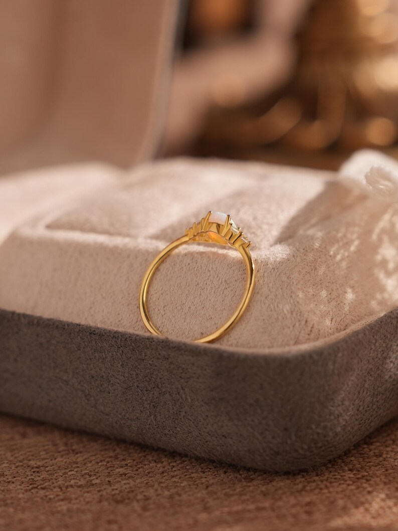 Puede incluir: Un anillo de oro con una gema rectangular blanquecina en el centro, flanqueada por piedras m&aacute;s peque&ntilde;as y transparentes. El anillo se presenta sobre una superficie gris y beige, mostrando su delicado dise&ntilde;o. La banda del anillo es fina y elegante.