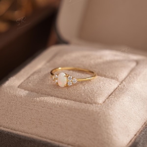 Puede incluir: Un anillo de oro con una piedra central de &oacute;palo ovalada y peque&ntilde;os diamantes. El anillo est&aacute; engastado en una banda delicada y se muestra en un joyero de terciopelo gris. Es una joya.