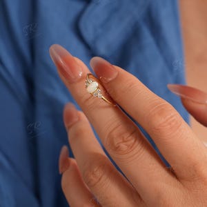 Puede incluir: Un anillo de oro con una piedra central de &oacute;palo ovalada, flanqueada por piedras de acento claras m&aacute;s peque&ntilde;as. El anillo est&aacute; engastado en una delicada banda de oro. El fondo es azul liso.