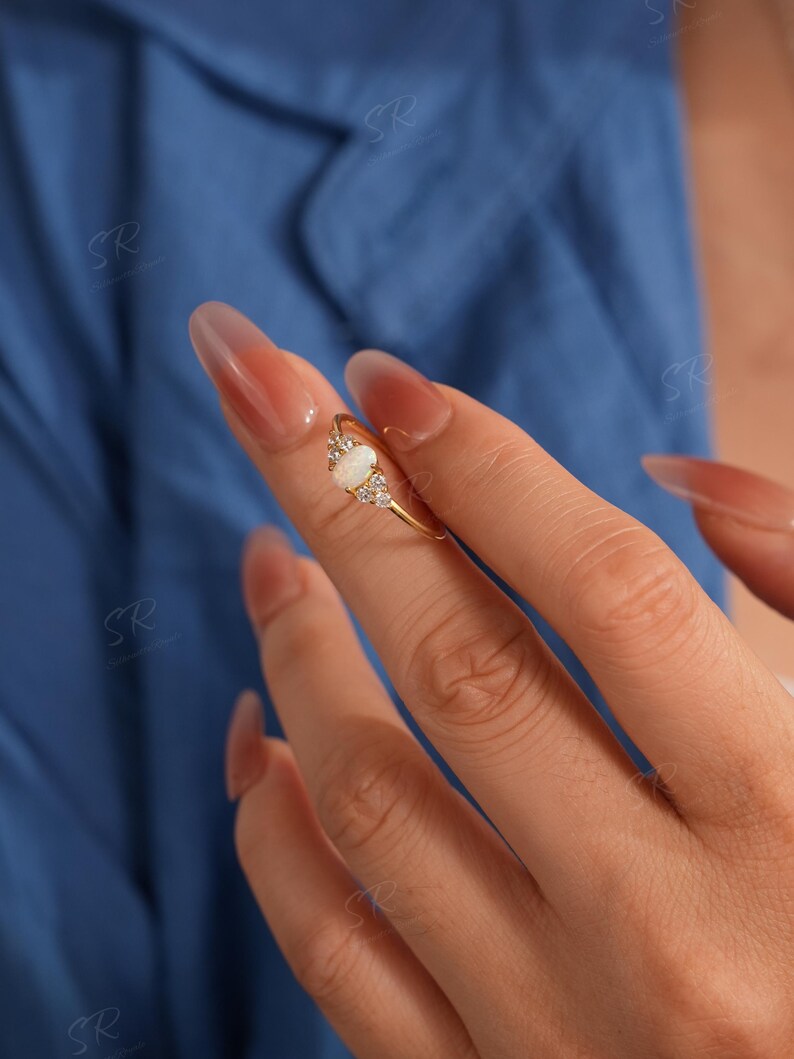 Puede incluir: Un anillo de oro con una piedra central de &oacute;palo ovalada, flanqueada por piedras de acento claras m&aacute;s peque&ntilde;as. El anillo est&aacute; engastado en una delicada banda de oro. El fondo es azul liso.